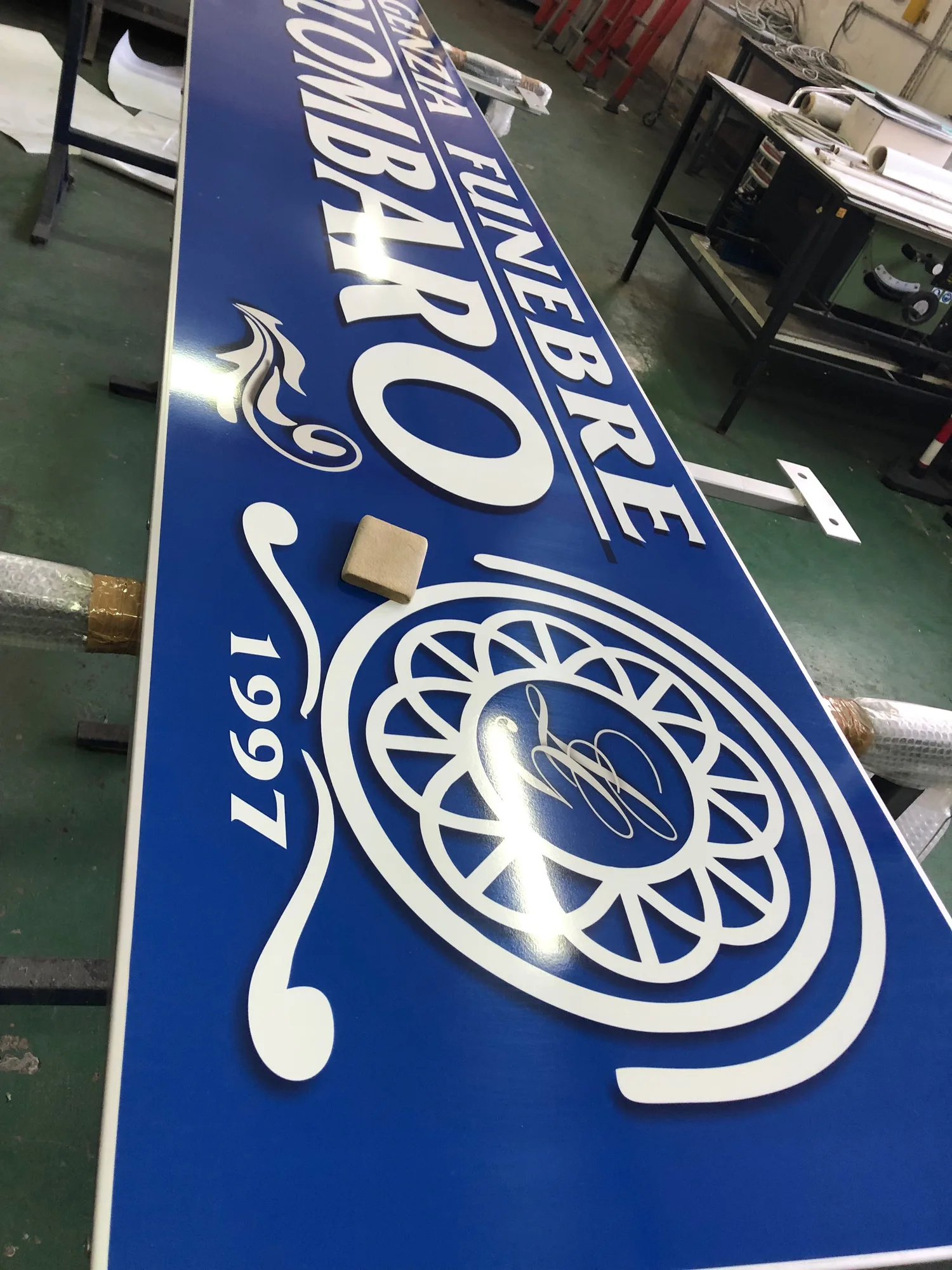 Grande insegna blu con scritte e decori bianchi in laboratorio, pronta per l'installazione