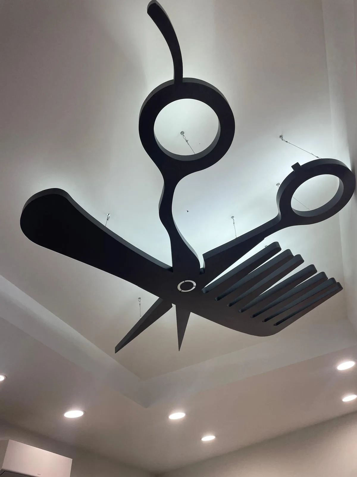 Grande sagoma di forbici e pettine appesa al soffitto di un salone, con luce sul retro