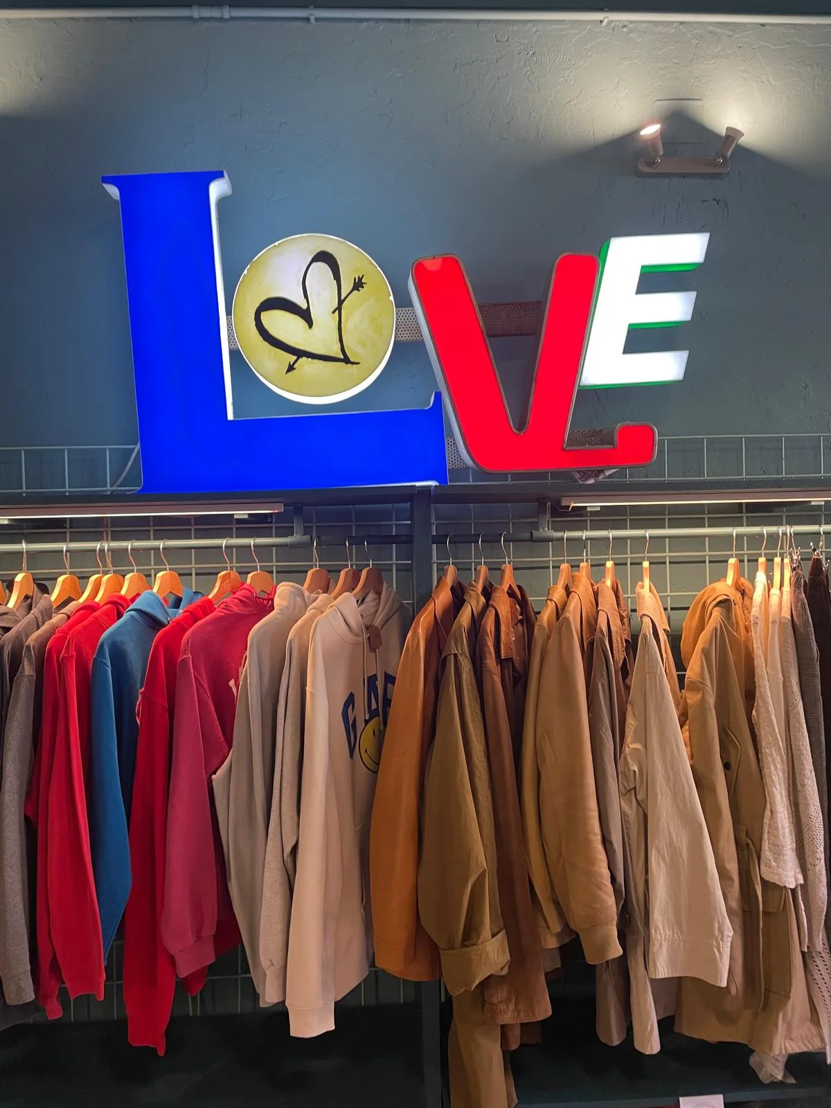 Parete di un negozio di abbigliamento con la scritta LOVE a lettere luminose blu, gialla, rossa e bianco-verde sopra gli appendiabiti